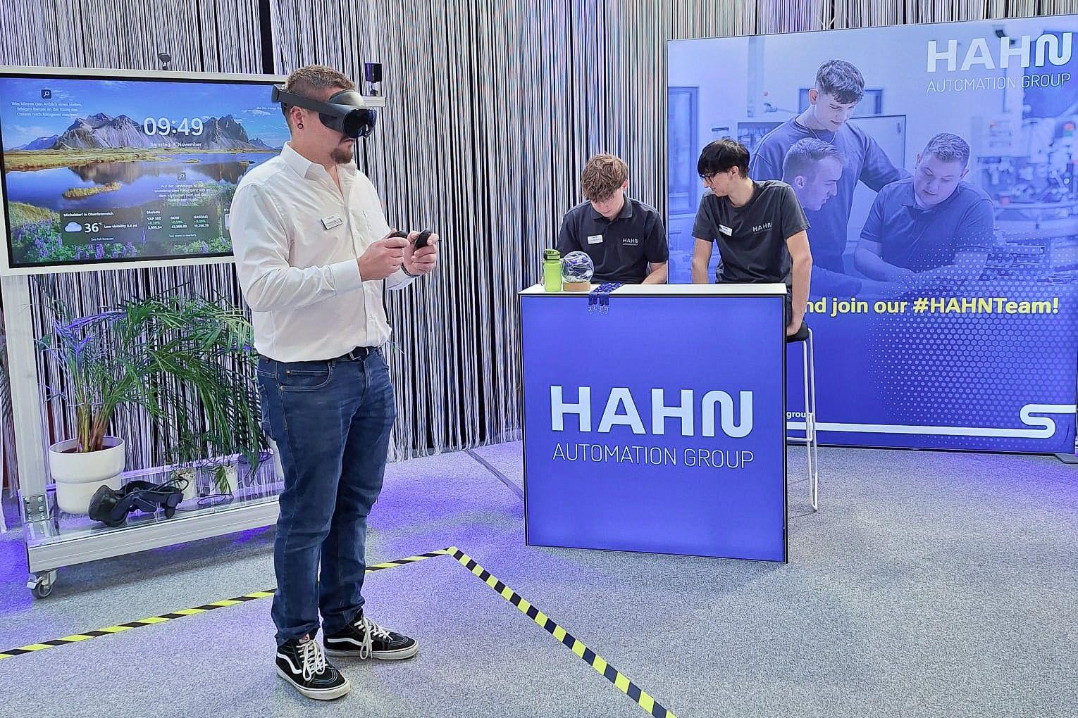 Zu sehen ist ein junger Mann, der mit VR-Brille vor einem Bildschirm navigiert, im Hintergrund zwei weitere junge Männer an einem mit Hahn gebrandeten Tresen.