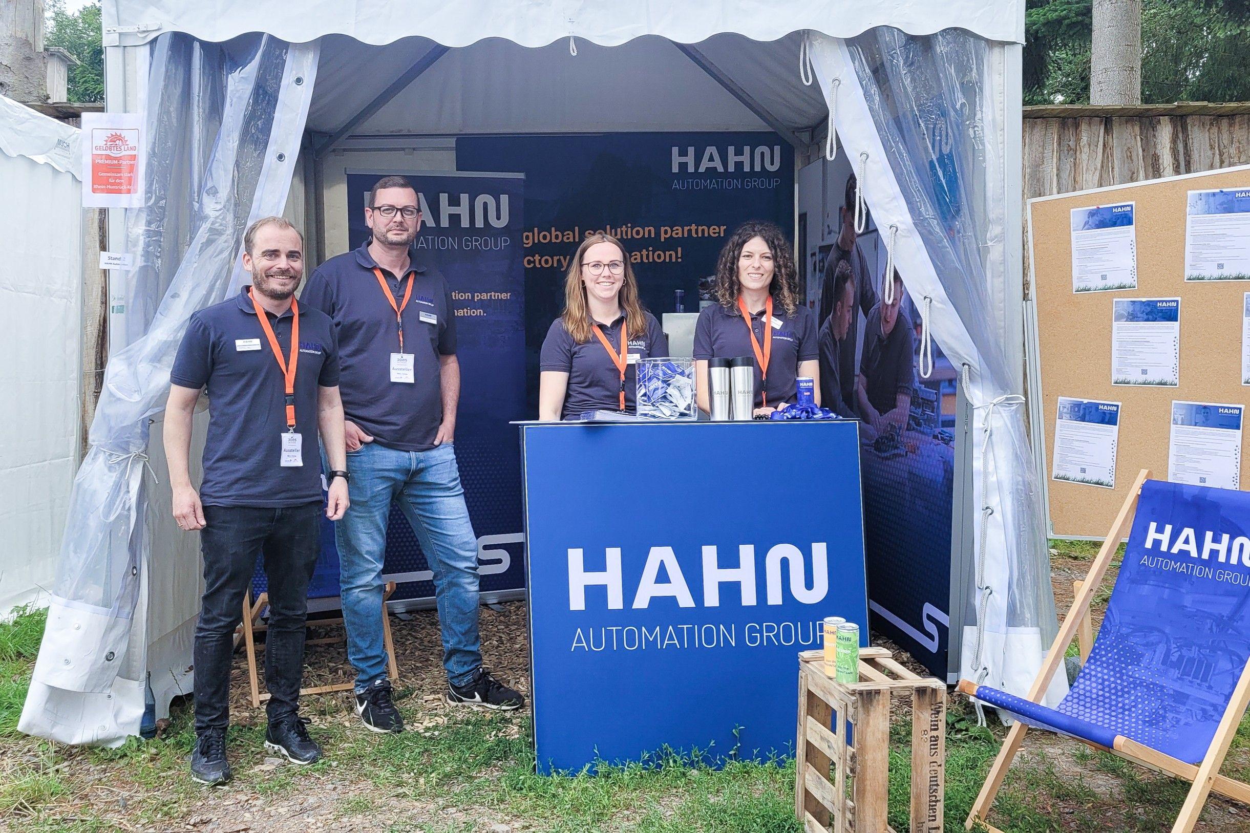Zwei junge Männer und Frauen in Hahn-Poloshirts sind vor einem Zelt im Grünen an einem mit Hahn gebrandeten Tresen zu sehen.
