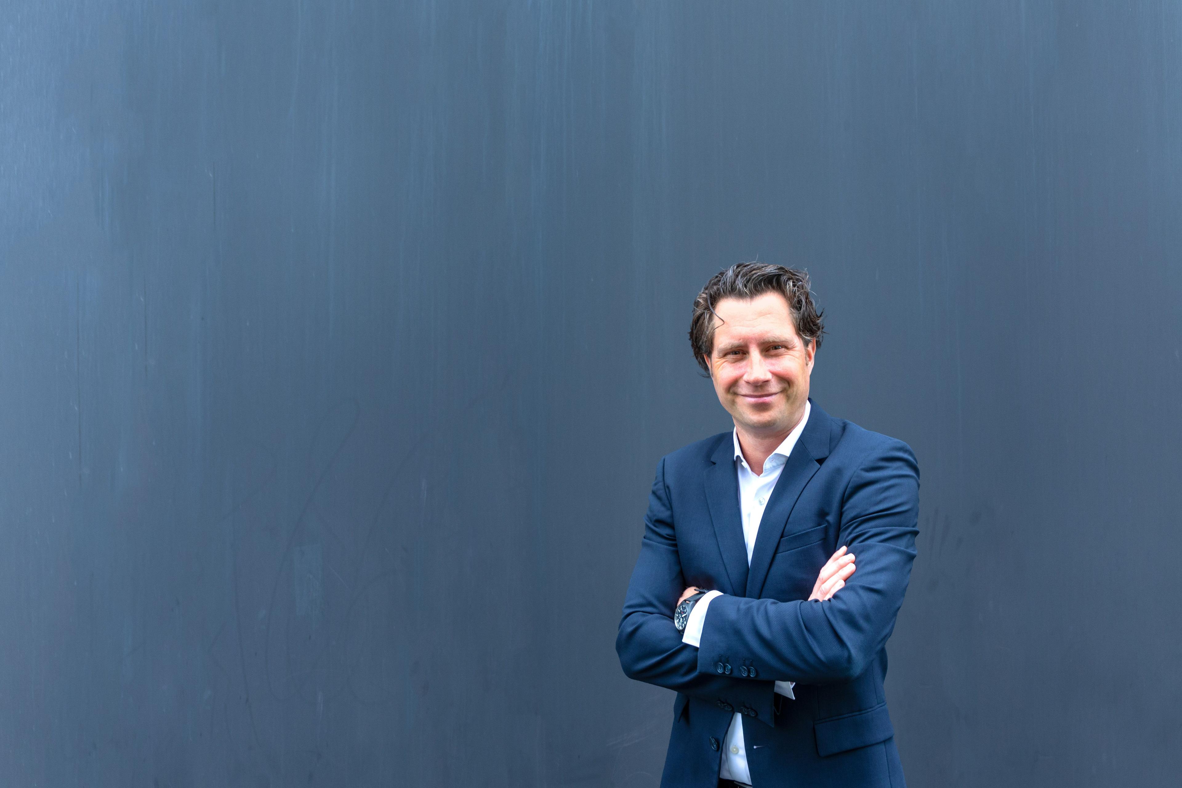 Christoph Tegeler in blauem Blazer mit verschränkten Armen schmunzelndvor einer blauen Wand.