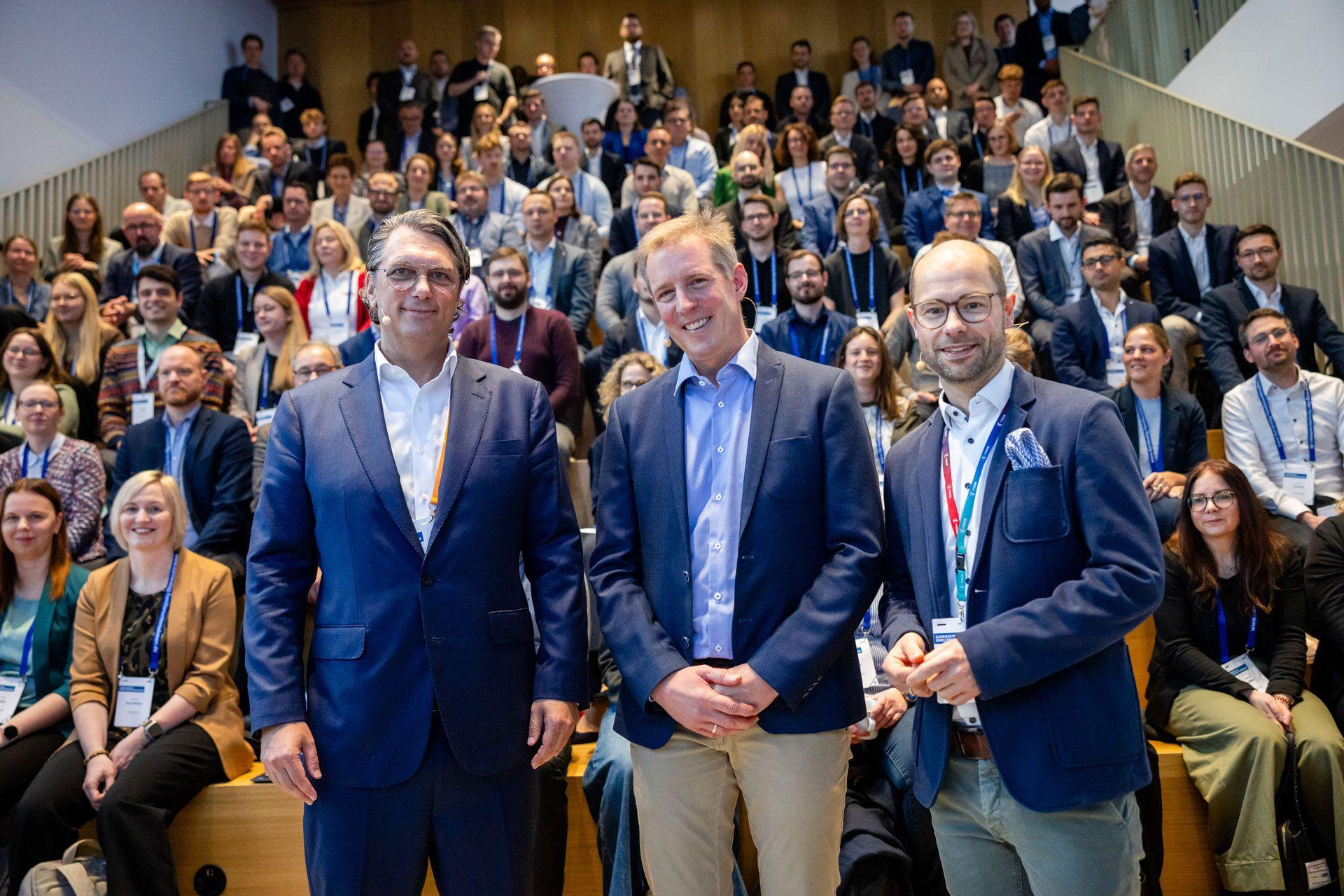 Drei Männer stehen vor einer Gruppe sitzender Frauen und Männer